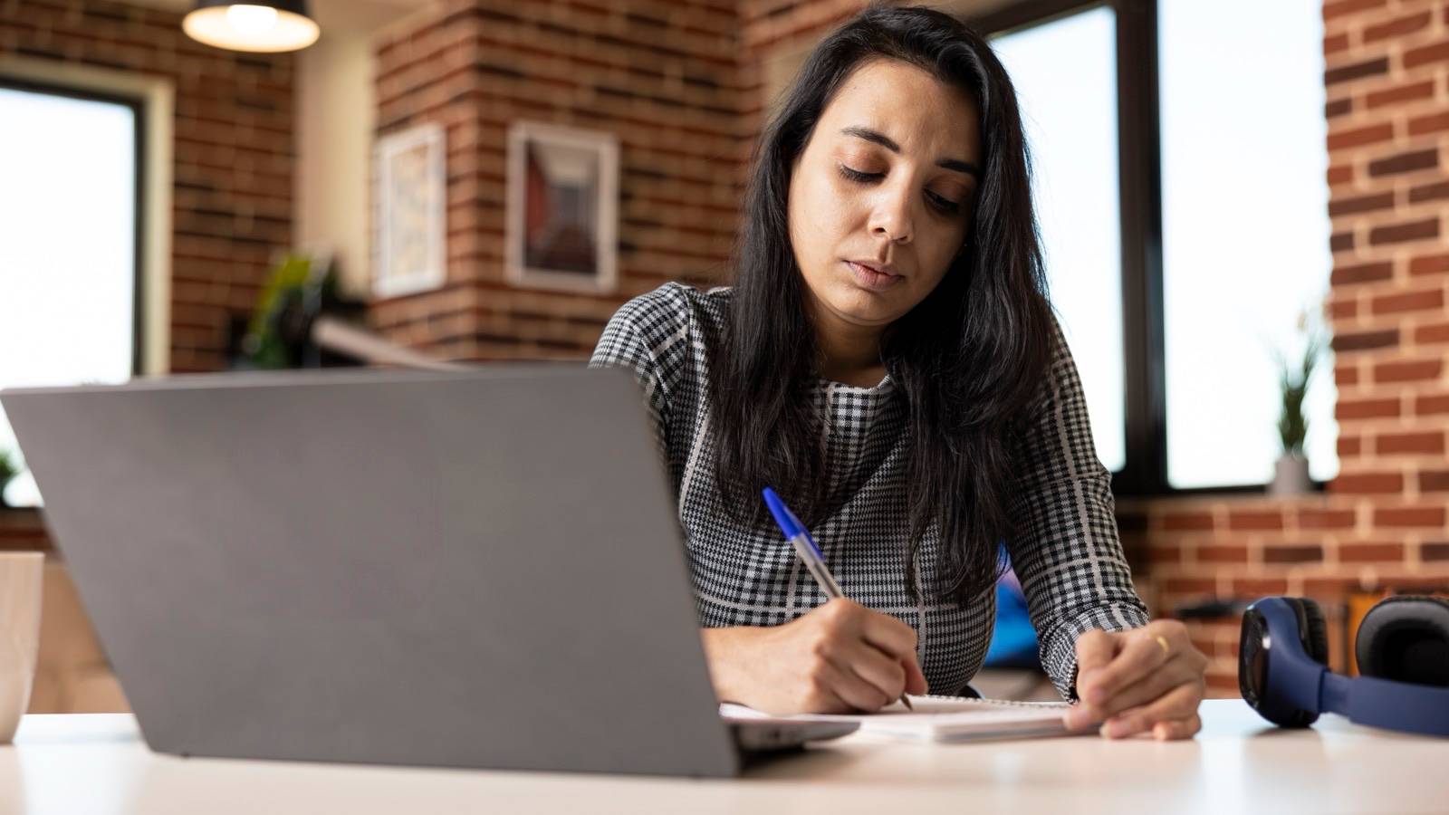 Woman taking notes beside her laptop at a desk
